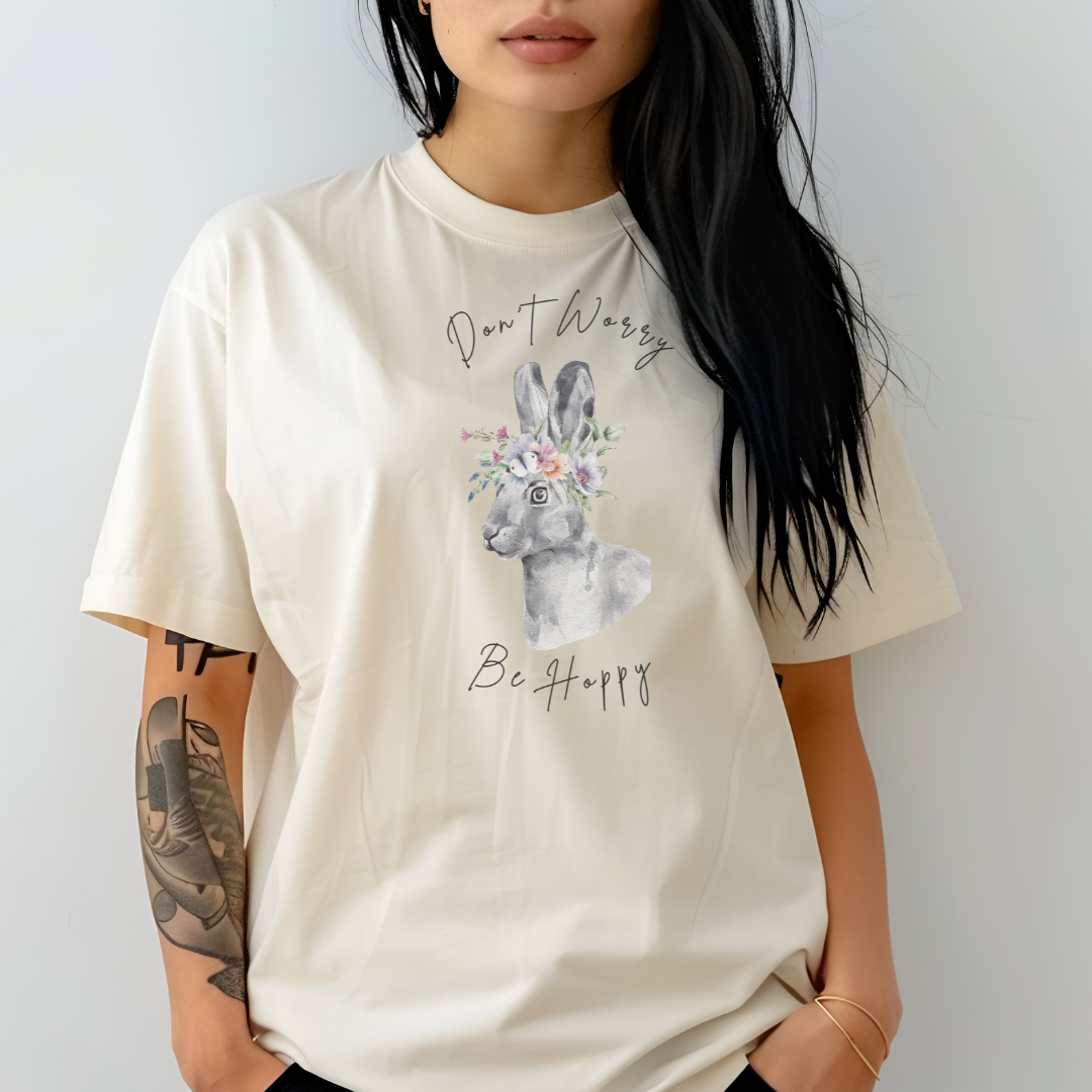 "Don't Worry, Be Hoppy!" Unisex Garment-Dyed T-Shirt, featuring a charming watercolor illustration of a grey rabbit wearing a crown of delicate pink and purple flowers, with the text "Don't Worry Be Hoppy" in a playful script above and below, modeled by a woman wearing size L. Color Ivory