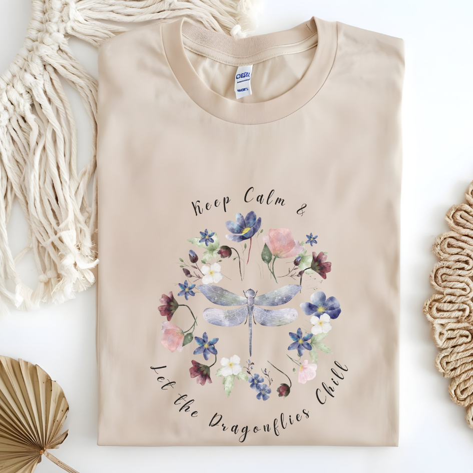 Flat lay of the "Chill Spring! Keep Calm & Let the Dragonflies Chill" T-Shirt, highlighting the soft, 100% ring-spun cotton fabric, classic crew neck, and the adorable dragonfly and floral wreath design. Color Sand