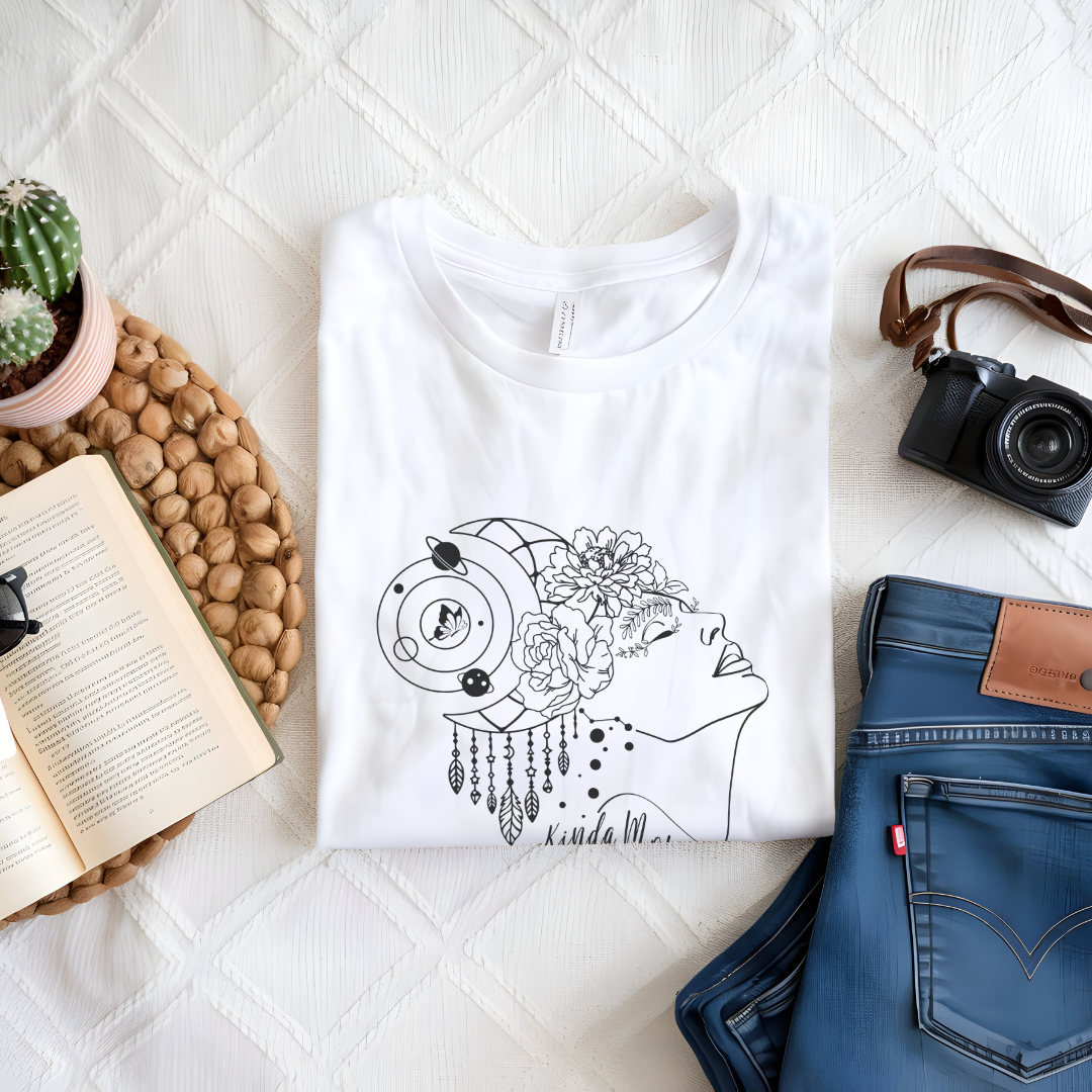 Close-up view of the "Kinda Mom" design on the Mother's Day Tee, showcasing the graphic of a minimalist line drawing of a woman's profile facing right., her hair is adorned with a crescent moon and dangling charms, giving a mystical or bohemian vibe. Overlaid on her hair is a cluster of blooming flowers, adding a touch of femininity and nature. Below the profile, in a casual, handwritten-style font, are the words "Kinda Mom." Color White