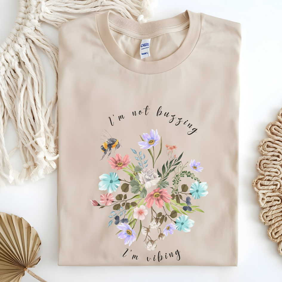 Flat lay of the "Sweet Spring! I'm Not Buzzing" T-Shirt, highlighting the soft, 100% ring-spun cotton fabric, classic crew neck, and the adorable bee and wildflower design. Color Sand