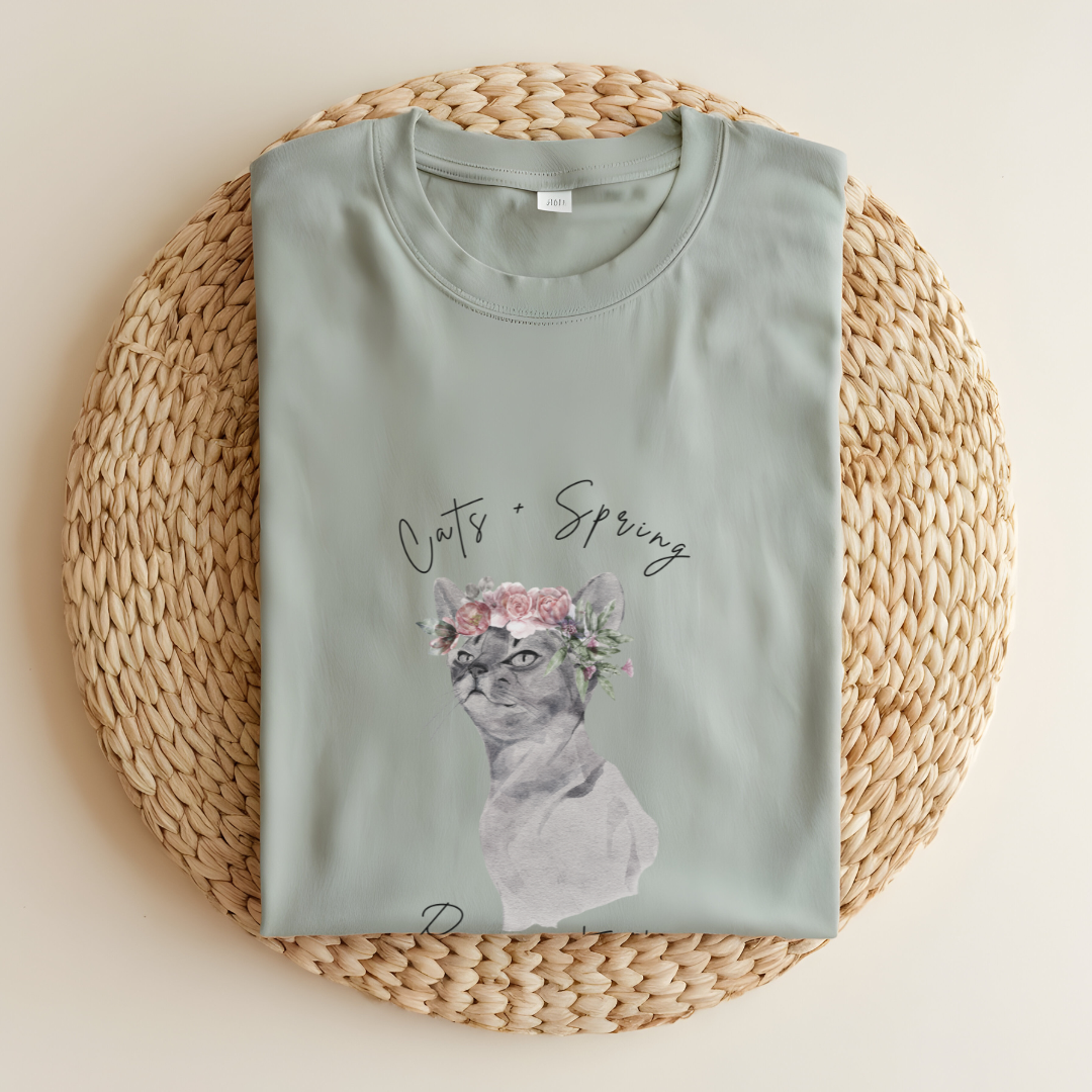 Flat lay of the "Purrfect Spring Vibes" T-Shirt, highlighting the garment-dyed fabric's texture and the durable double-needle stitching. The watercolor cat and floral crown design. Color Bay