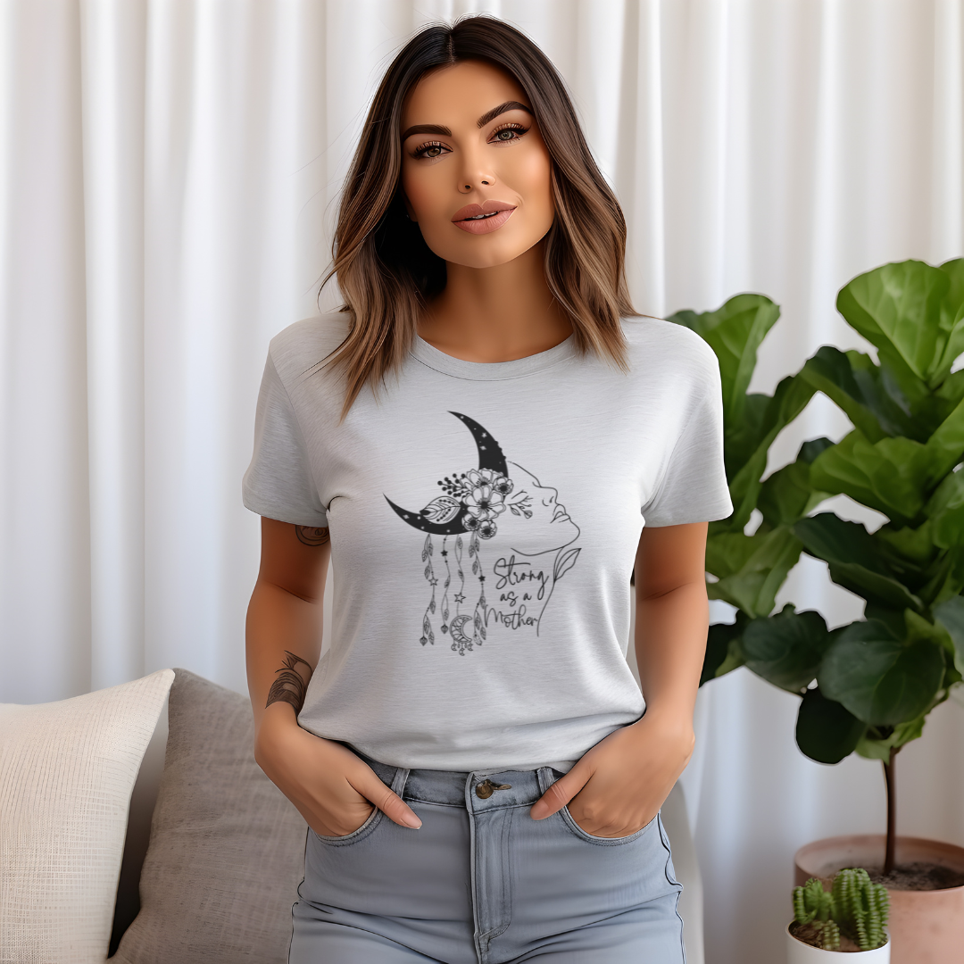 "Strong as a Mother" Unisex Jersey T-Shirt, featuring a powerful graphic of a woman's profile emerging from a crescent moon, adorned with blooming flowers and delicate dangling charms, modeled by a Woman wearing size M. Color Ash