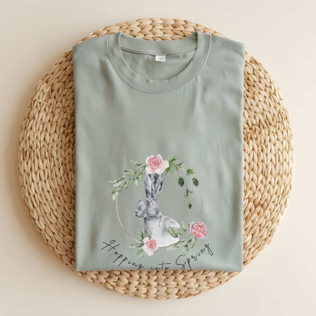 Flat lay of the "Hoppin' into Spring" T-Shirt, highlighting the garment-dyed fabric's texture and the durable double-needle stitching. The watercolor bunny and floral wreath design are clearly visible. ColorBay