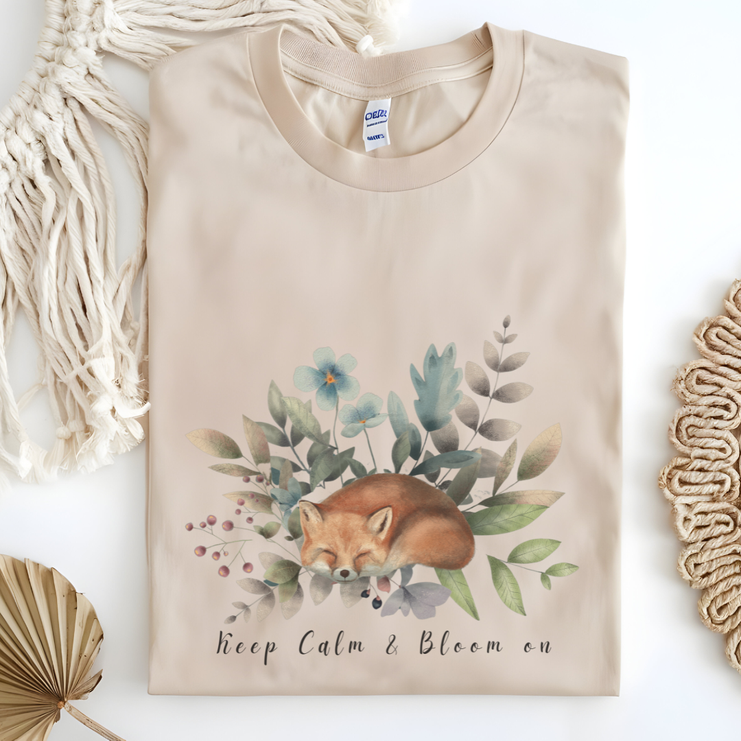 Flat lay of the "Keep Calm & Bloom On" T-Shirt, highlighting the soft, 100% US cotton fabric and classic crew neck. The watercolor fox and floral design are clearly visible. Color Sand