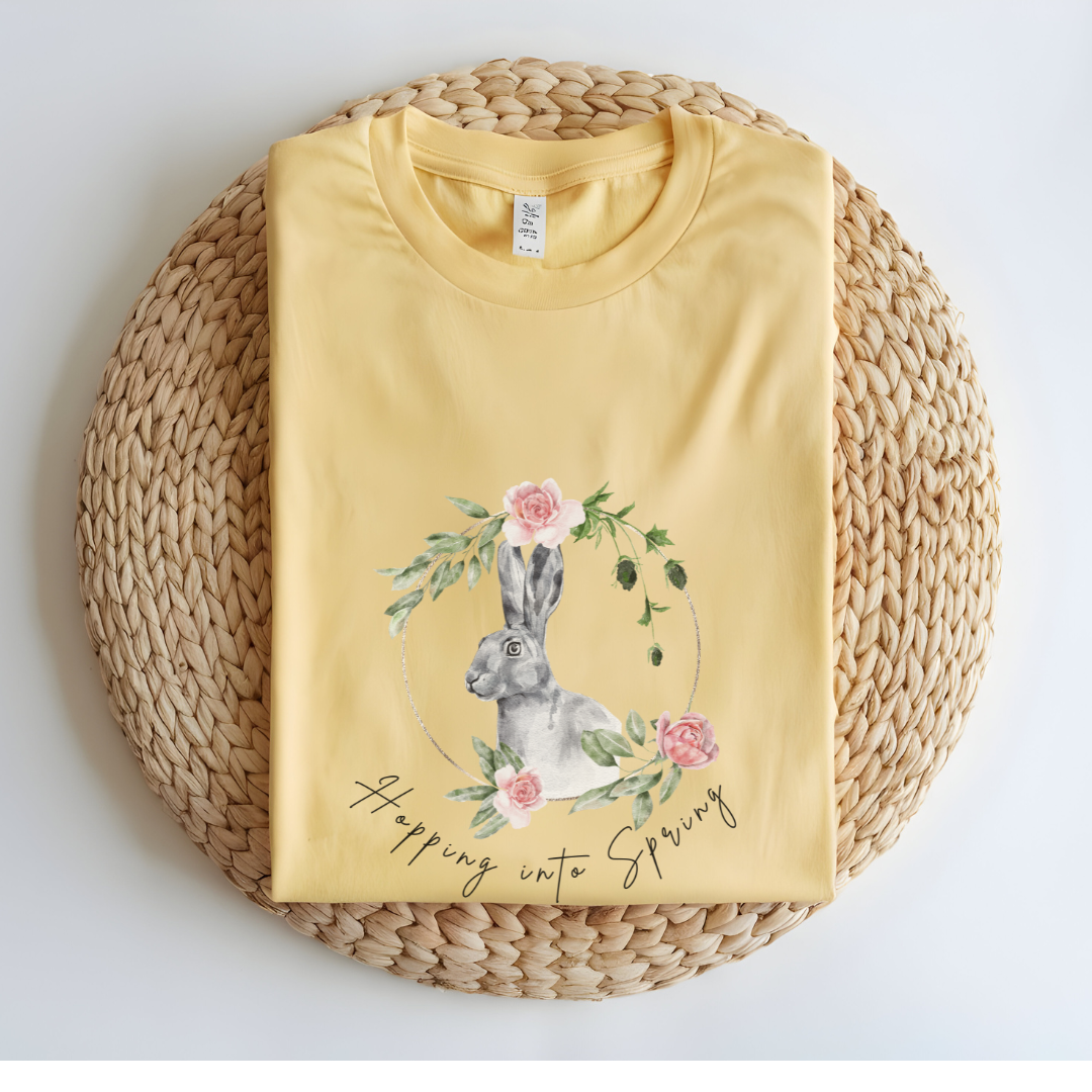 Flat lay of the "Hoppin' into Spring" T-Shirt, highlighting the garment-dyed fabric's texture and the durable double-needle stitching. The watercolor bunny and floral wreath design are clearly visible. Color Butter