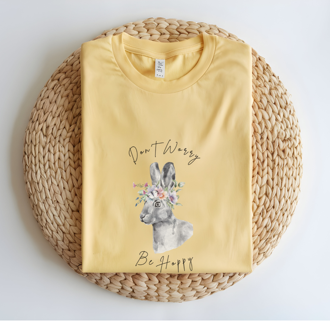 Flat lay of the "Don't Worry, Be Hoppy!" T-Shirt, highlighting the garment-dyed fabric's texture and the durable double-needle stitching. The watercolor bunny and floral crown design are clearly visible. Color Butter