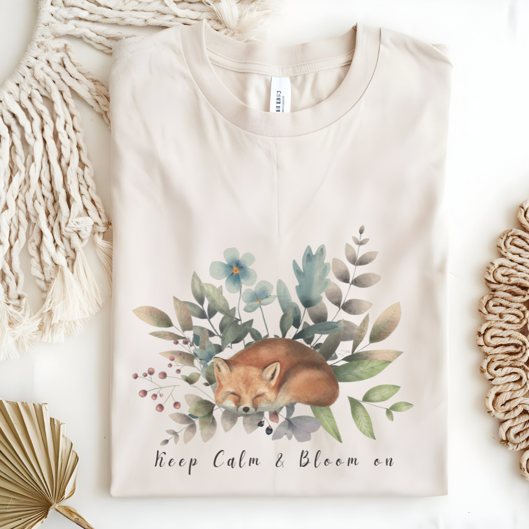 Flat lay of the "Keep Calm & Bloom On" T-Shirt, highlighting the soft, 100% US cotton fabric and classic crew neck. The watercolor fox and floral design are clearly visible. Color Natural