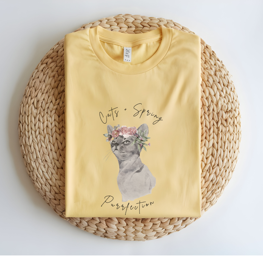 Flat lay of the "Purrfect Spring Vibes" T-Shirt, highlighting the garment-dyed fabric's texture and the durable double-needle stitching. The watercolor cat and floral crown design. Color Butter