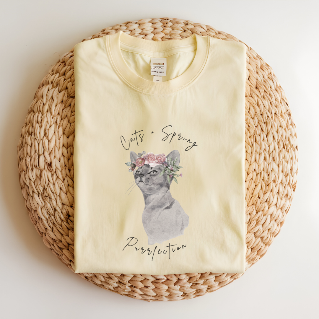 Flat lay of the "Purrfect Spring Vibes" T-Shirt, highlighting the garment-dyed fabric's texture and the durable double-needle stitching. The watercolor cat and floral crown design. Color Banana