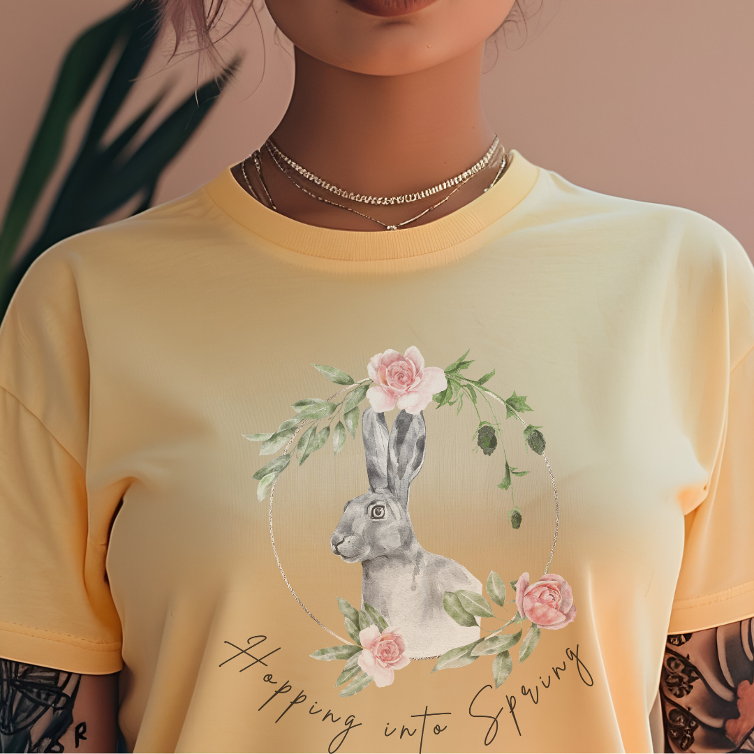 "Don't Worry, Be Hoppy!" Unisex Garment-Dyed T-Shirt, featuring a charming watercolor illustration of a grey rabbit wearing a crown of delicate pink and purple flowers, with the text "Don't Worry Be Hoppy" in a playful script above and below, modeled by a woman wearing size L. Color Banana