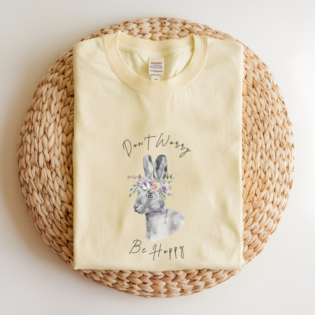 Flat lay of the "Don't Worry, Be Hoppy!" T-Shirt, highlighting the garment-dyed fabric's texture and the durable double-needle stitching. The watercolor bunny and floral crown design are clearly visible. Color Banana