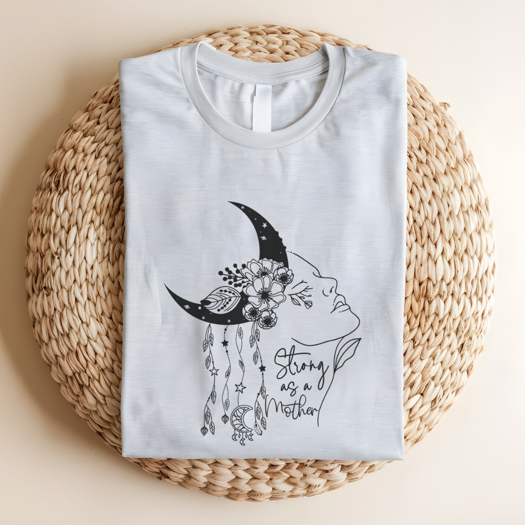 Close-up view of the "Strong as a Mother" graphic, showcasing the intricate details of the crescent moon with its stippled texture, the vibrant flowers nestled within it, and the flowing lines of the woman's profile. The dangling charms add a touch of whimsy. The words "Strong as a Mother" are written in a delicate script, font beneath the graphic. Color Ash