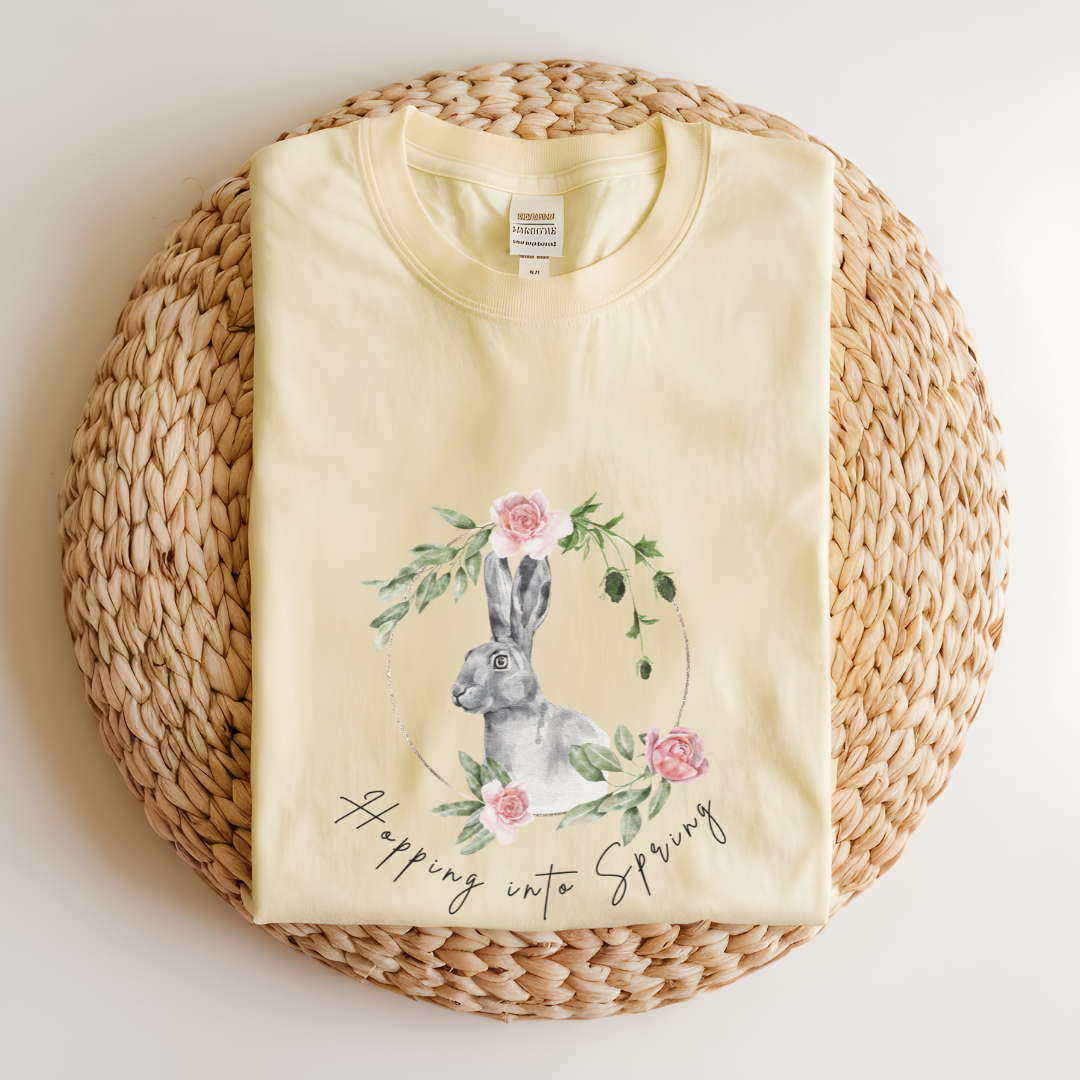 Flat lay of the "Hoppin' into Spring" T-Shirt, highlighting the garment-dyed fabric's texture and the durable double-needle stitching. The watercolor bunny and floral wreath design are clearly visible. Color Banana