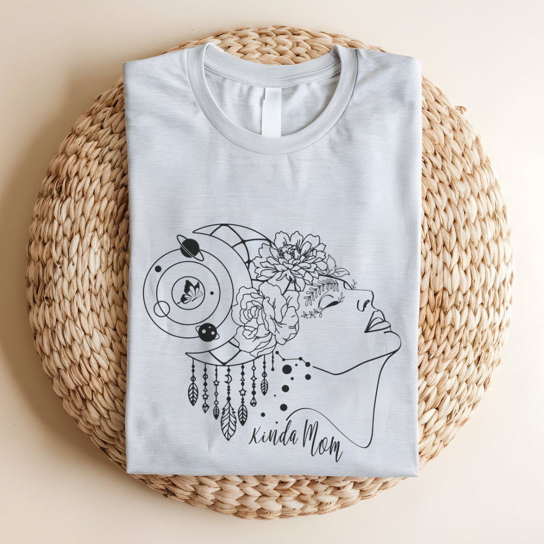 Close-up view of the "Kinda Mom" design on the Mother's Day Tee, showcasing the graphic of a minimalist line drawing of a woman's profile facing right., her hair is adorned with a crescent moon and dangling charms, giving a mystical or bohemian vibe. Overlaid on her hair is a cluster of blooming flowers, adding a touch of femininity and nature. Below the profile, in a casual, handwritten-style font, are the words "Kinda Mom." Color Ash