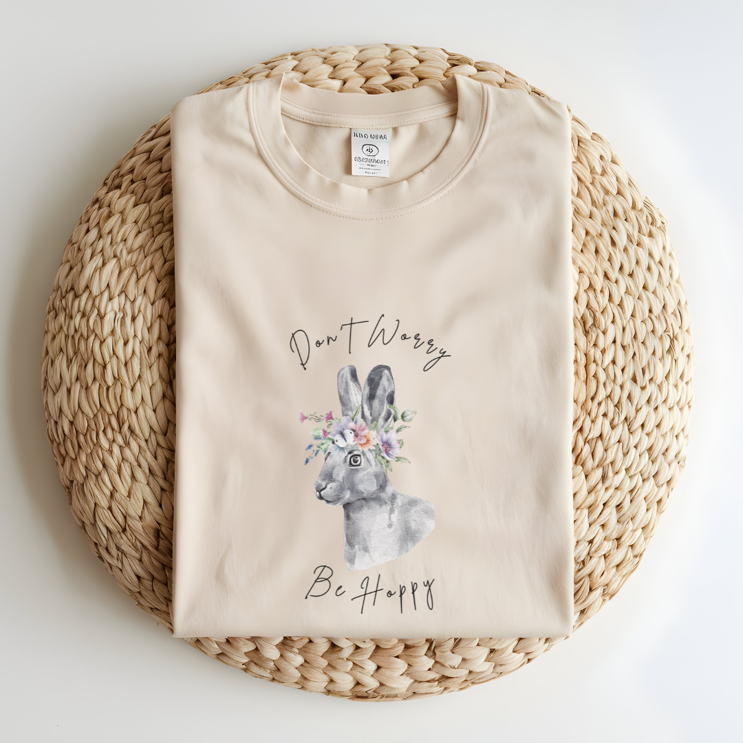 Flat lay of the "Don't Worry, Be Hoppy!" T-Shirt, highlighting the garment-dyed fabric's texture and the durable double-needle stitching. The watercolor bunny and floral crown design are clearly visible. Color Ivory