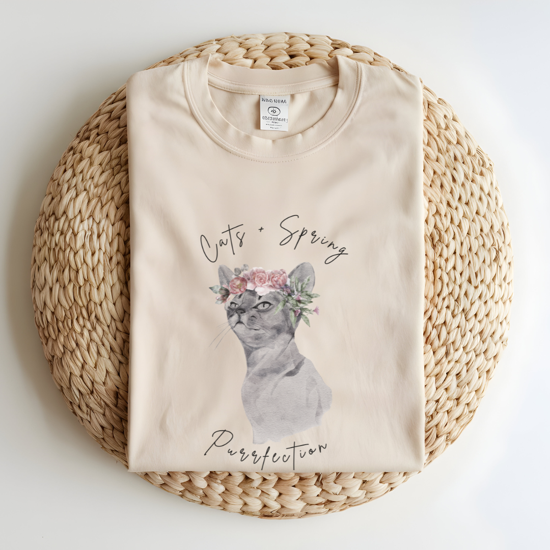 Flat lay of the "Purrfect Spring Vibes" T-Shirt, highlighting the garment-dyed fabric's texture and the durable double-needle stitching. The watercolor cat and floral crown design, color Ivory