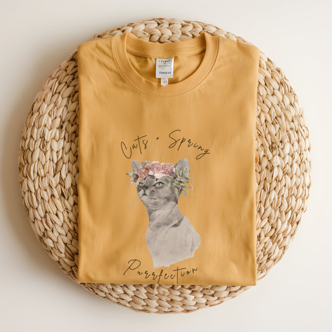 Flat lay of the "Purrfect Spring Vibes" T-Shirt, highlighting the garment-dyed fabric's texture and the durable double-needle stitching. The watercolor cat and floral crown design. Color Mustard