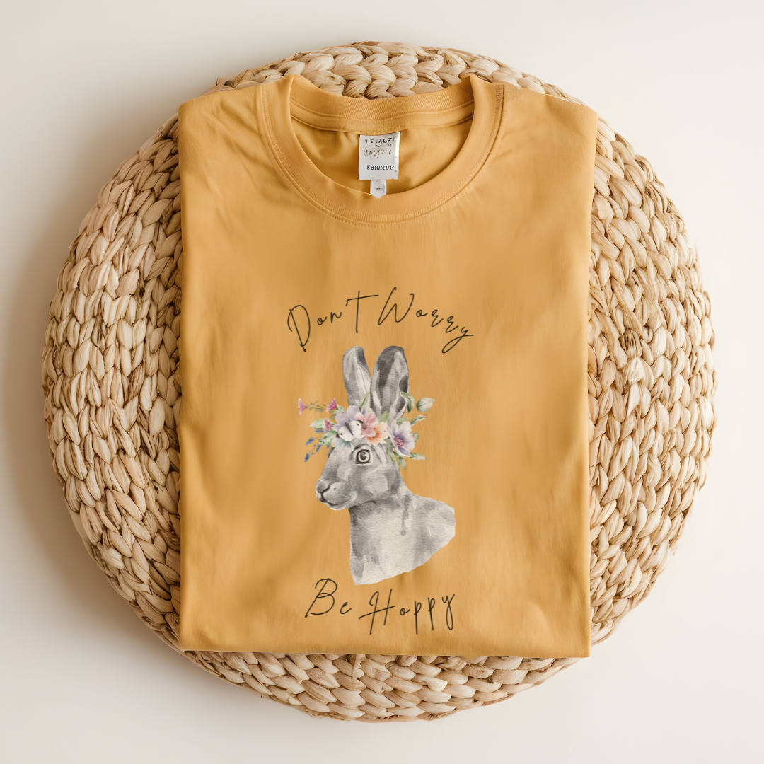 Flat lay of the "Don't Worry, Be Hoppy!" T-Shirt, highlighting the garment-dyed fabric's texture and the durable double-needle stitching. The watercolor bunny and floral crown design are clearly visible. Color Mustard