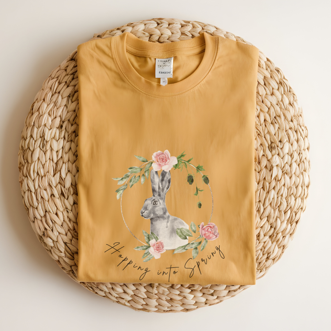 Flat lay of the "Hoppin' into Spring" T-Shirt, highlighting the garment-dyed fabric's texture and the durable double-needle stitching. The watercolor bunny and floral wreath design are clearly visible. Color Mustard