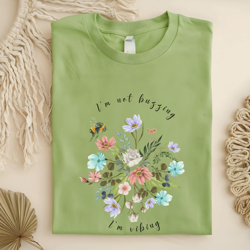 Flat lay of the "Sweet Spring! I'm Not Buzzing" T-Shirt, highlighting the soft, 100% ring-spun cotton fabric, classic crew neck, and the adorable bee and wildflower design. Color Pistachio