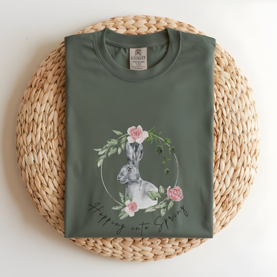 Flat lay of the "Hoppin' into Spring" T-Shirt, highlighting the garment-dyed fabric's texture and the durable double-needle stitching. The watercolor bunny and floral wreath design are clearly visible. Color Light green