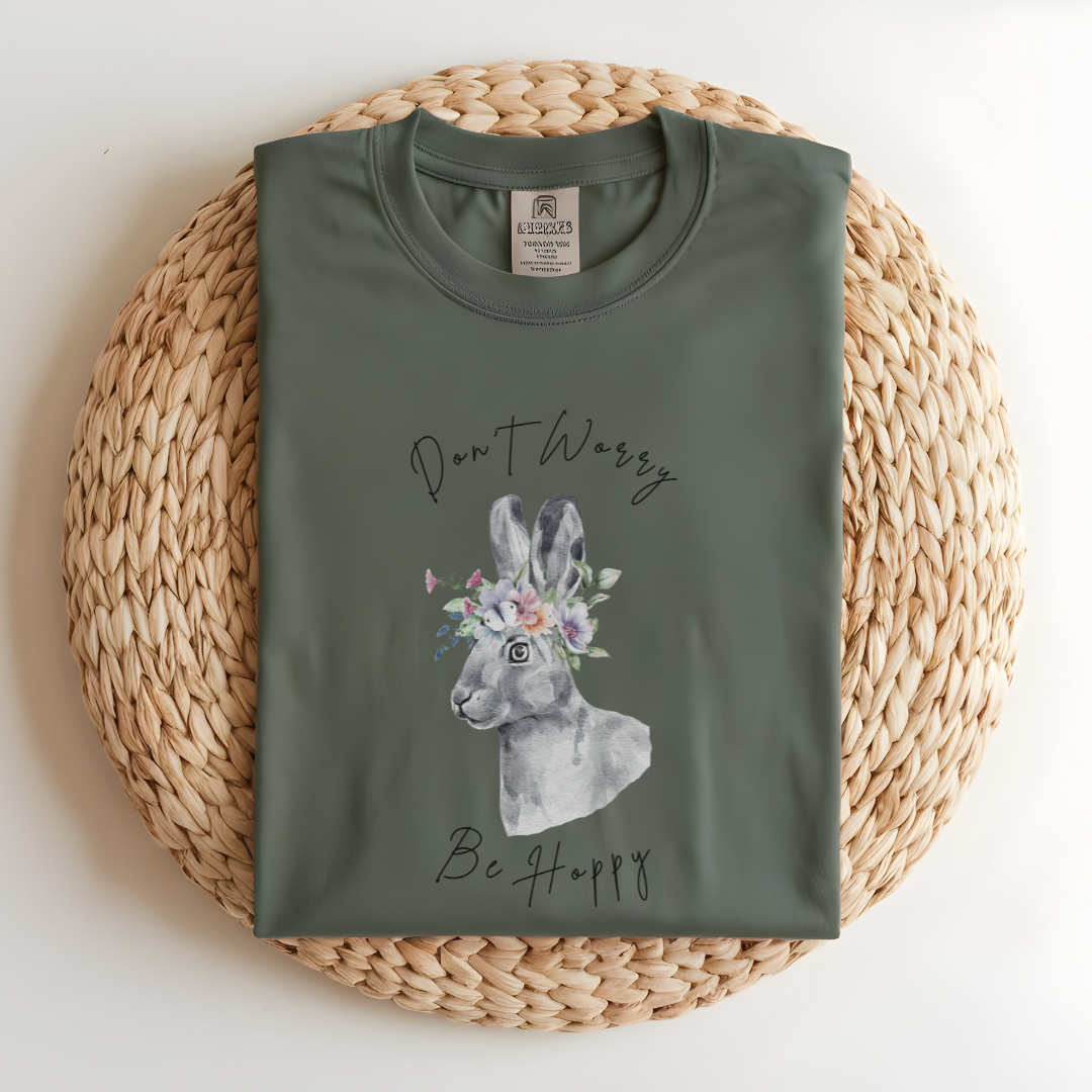 Flat lay of the "Don't Worry, Be Hoppy!" T-Shirt, highlighting the garment-dyed fabric's texture and the durable double-needle stitching. The watercolor bunny and floral crown design are clearly visible. Color Light green