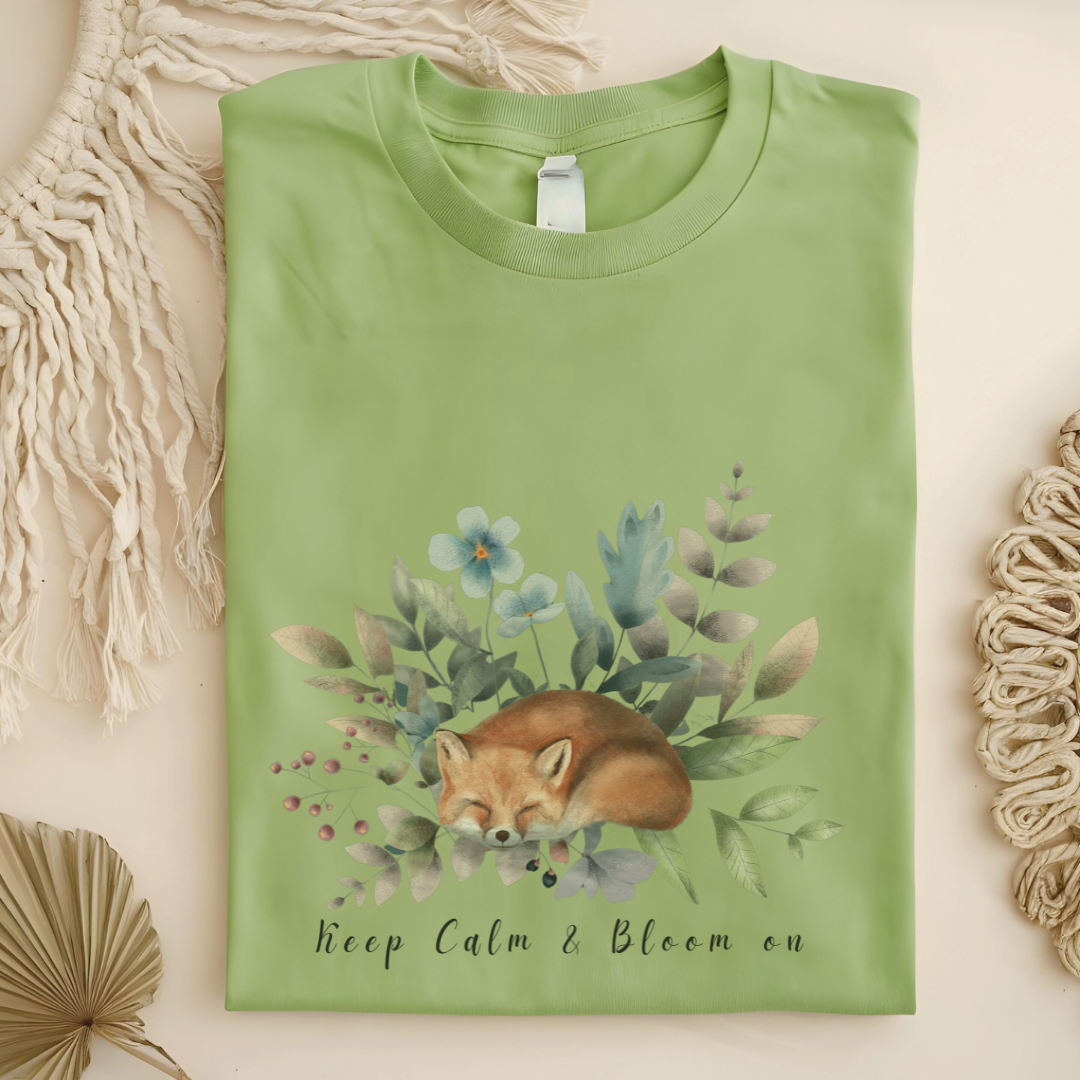 Flat lay of the "Keep Calm & Bloom On" T-Shirt, highlighting the soft, 100% US cotton fabric and classic crew neck. The watercolor fox and floral design are clearly visible. Color Pistaccio