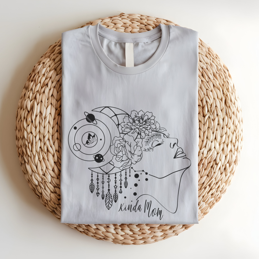 Close-up view of the "Kinda Mom" design on the Mother's Day Tee, showcasing the graphic of a minimalist line drawing of a woman's profile facing right., her hair is adorned with a crescent moon and dangling charms, giving a mystical or bohemian vibe. Overlaid on her hair is a cluster of blooming flowers, adding a touch of femininity and nature. Below the profile, in a casual, handwritten-style font, are the words "Kinda Mom." Color Heather Clay Grey