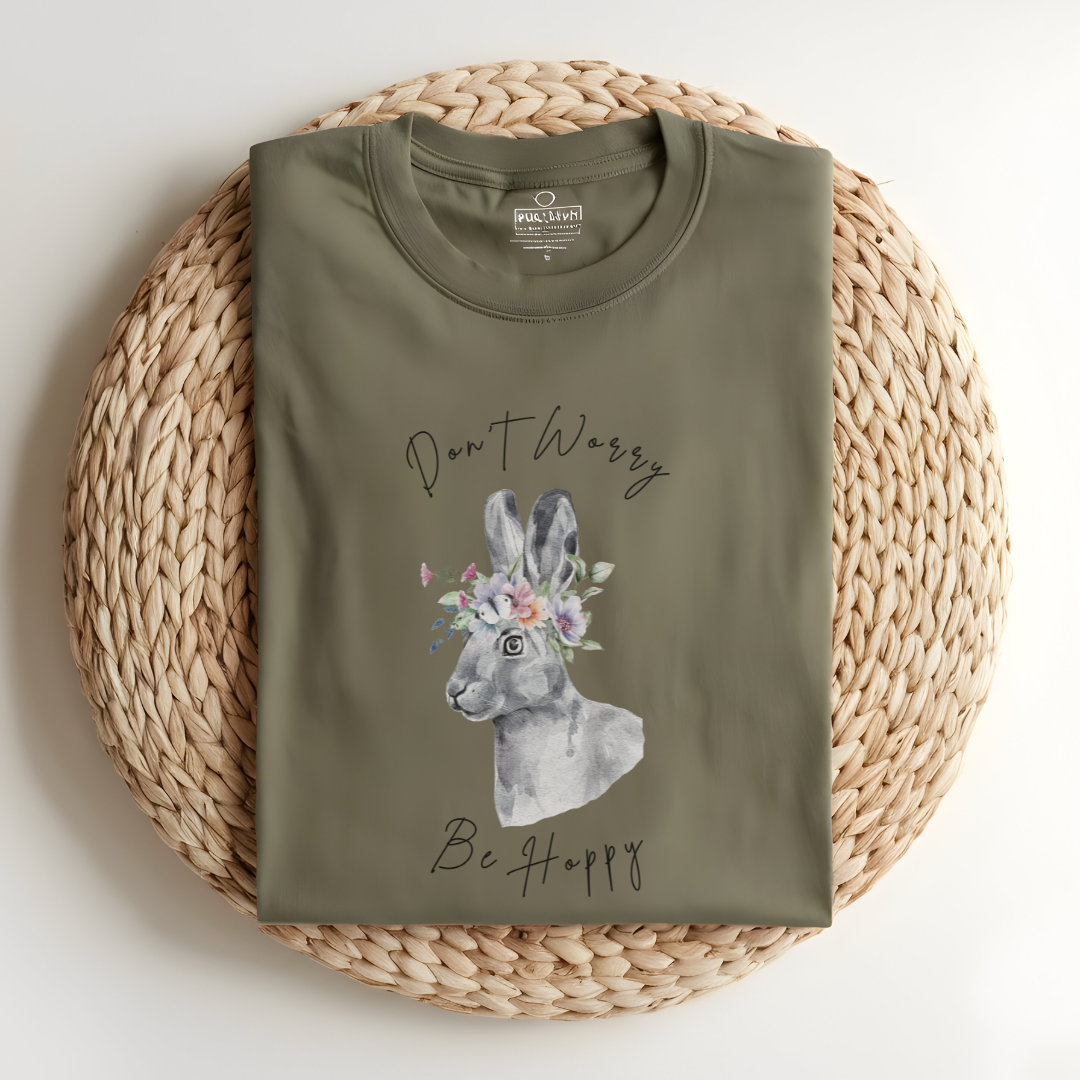 Flat lay of the "Don't Worry, Be Hoppy!" T-Shirt, highlighting the garment-dyed fabric's texture and the durable double-needle stitching. The watercolor bunny and floral crown design are clearly visible. Color Khaki