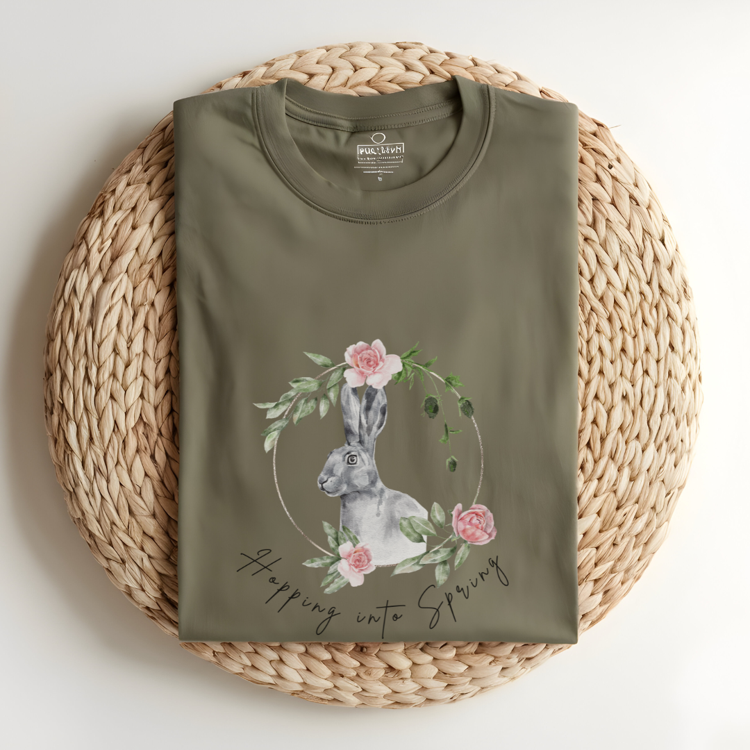 Flat lay of the "Hoppin' into Spring" T-Shirt, highlighting the garment-dyed fabric's texture and the durable double-needle stitching. The watercolor bunny and floral wreath design are clearly visible. Color Khaki