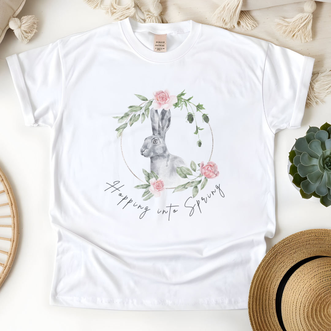 Flat lay of the "Hoppin' into Spring" T-Shirt, highlighting the garment-dyed fabric's texture and the durable double-needle stitching. The watercolor bunny and floral wreath design are clearly visible. Color White