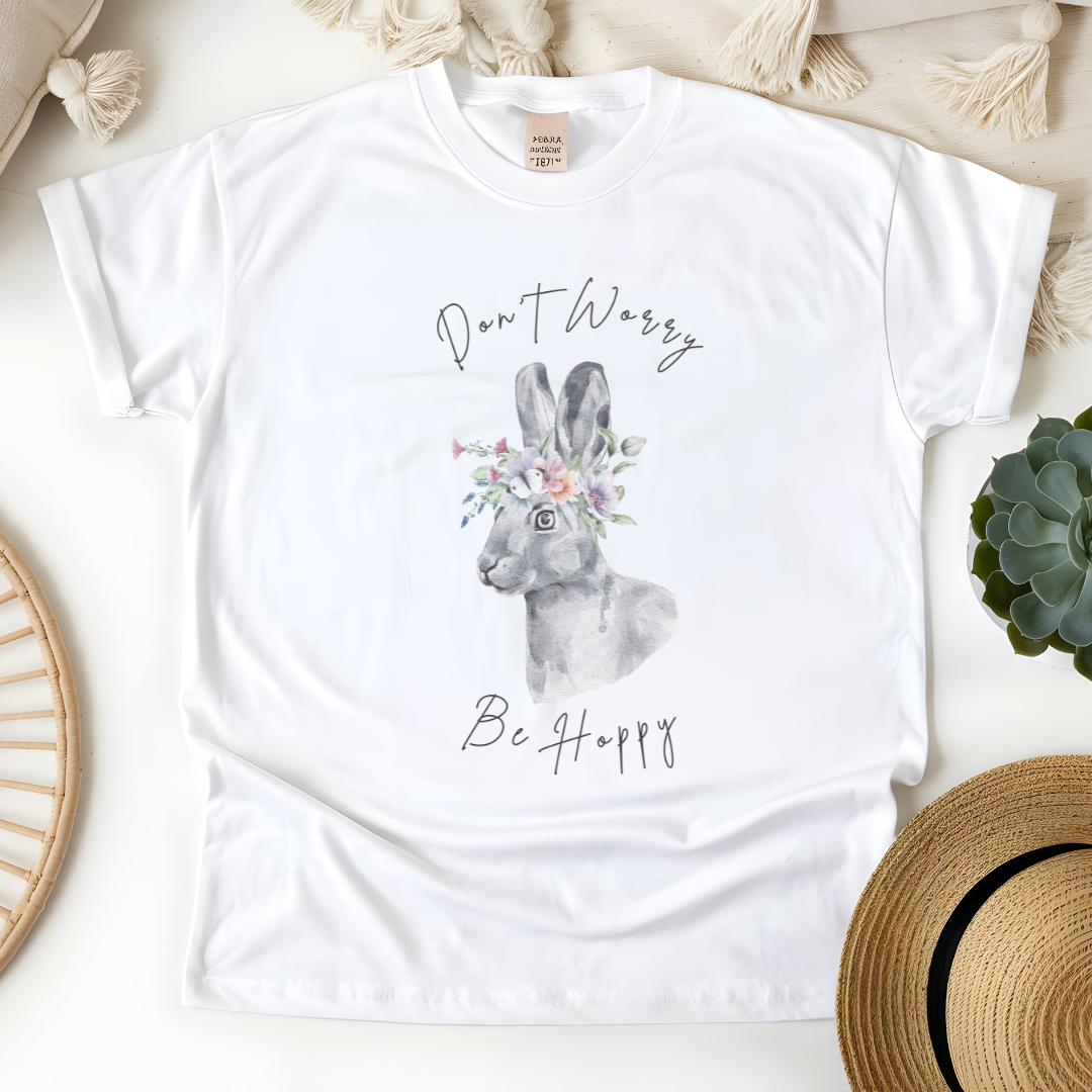 Flat lay of the "Don't Worry, Be Hoppy!" T-Shirt, highlighting the garment-dyed fabric's texture and the durable double-needle stitching. The watercolor bunny and floral crown design are clearly visible. Color White