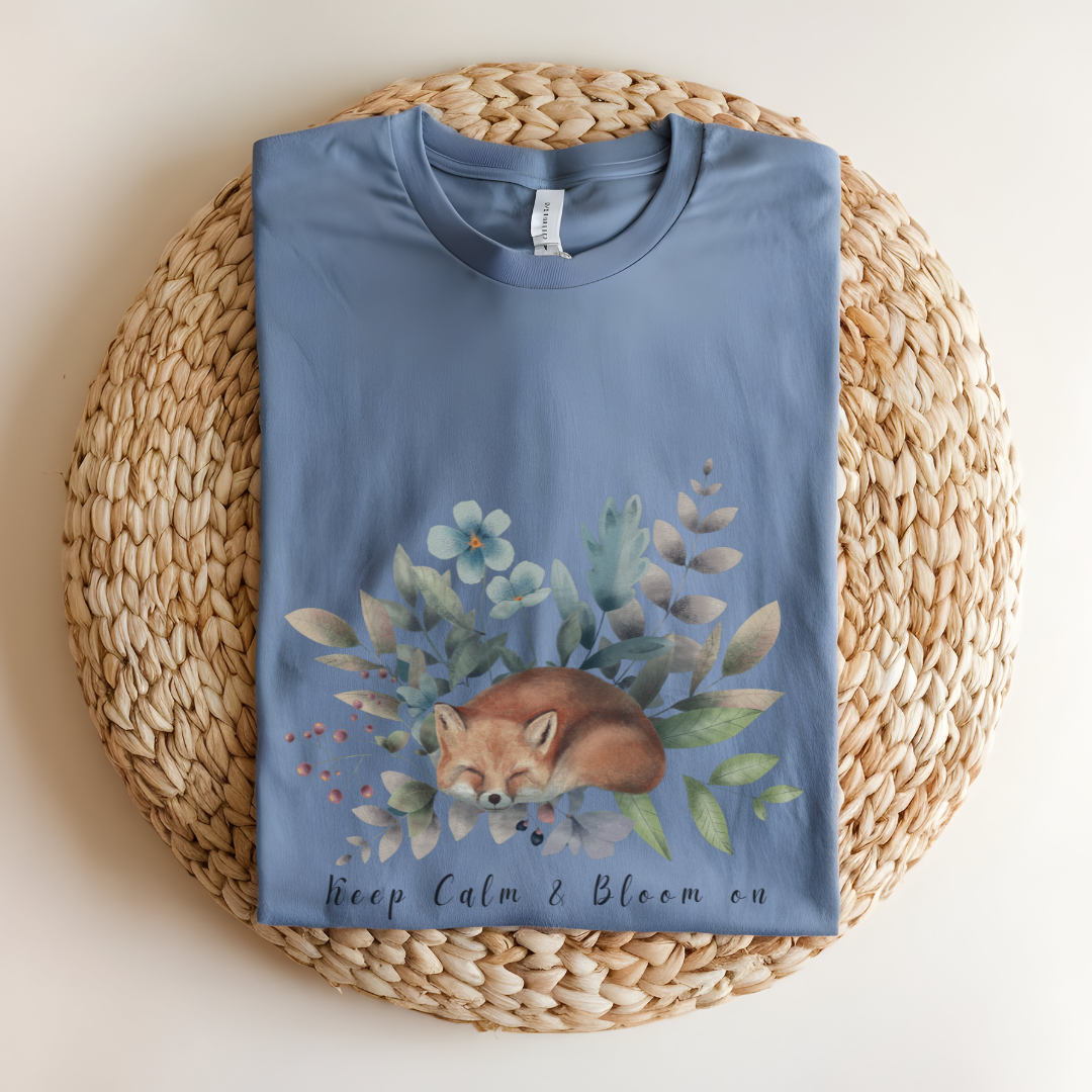 Flat lay of the "Keep Calm & Bloom On" T-Shirt, highlighting the soft, 100% US cotton fabric and classic crew neck. The watercolor fox and floral design are clearly visible. Color Heather indigo