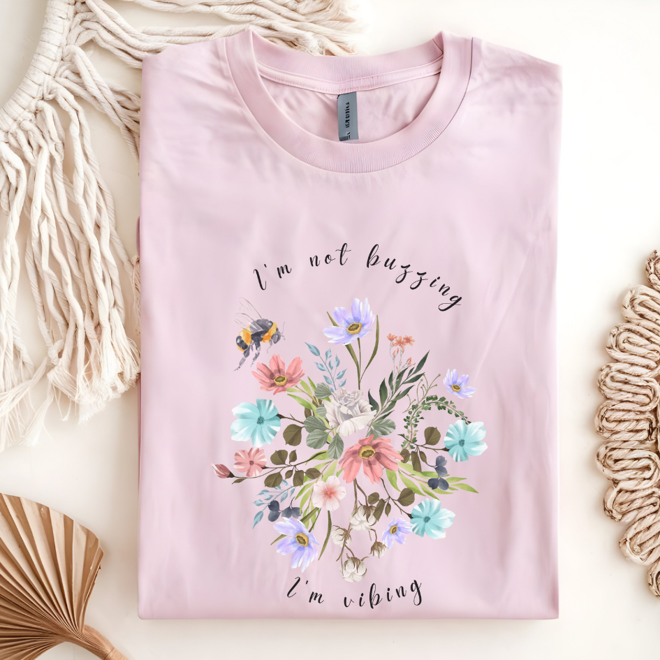 Flat lay of the "Sweet Spring! I'm Not Buzzing" T-Shirt, highlighting the soft, 100% ring-spun cotton fabric, classic crew neck, and the adorable bee and wildflower design. Color Light pink