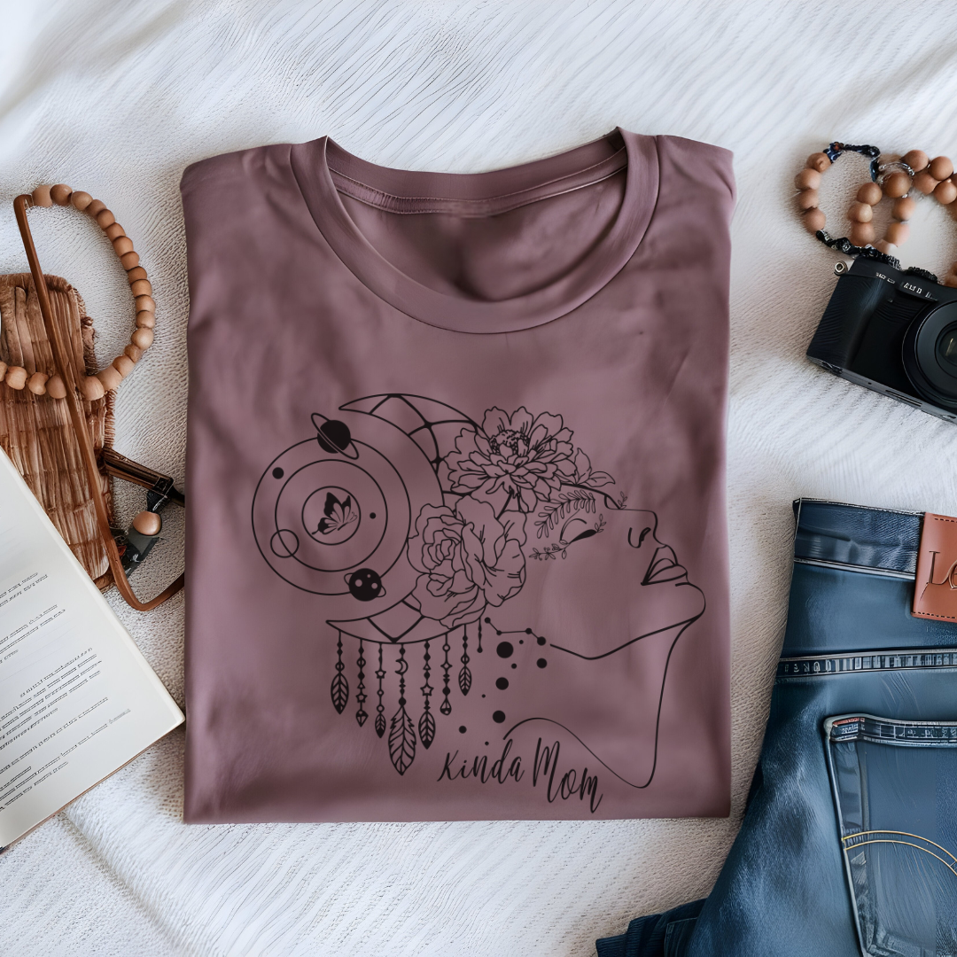 Close-up view of the "Kinda Mom" design on the Mother's Day Tee, showcasing the graphic of a minimalist line drawing of a woman's profile facing right., her hair is adorned with a crescent moon and dangling charms, giving a mystical or bohemian vibe. Overlaid on her hair is a cluster of blooming flowers, adding a touch of femininity and nature. Below the profile, in a casual, handwritten-style font, are the words "Kinda Mom." Color Mauve