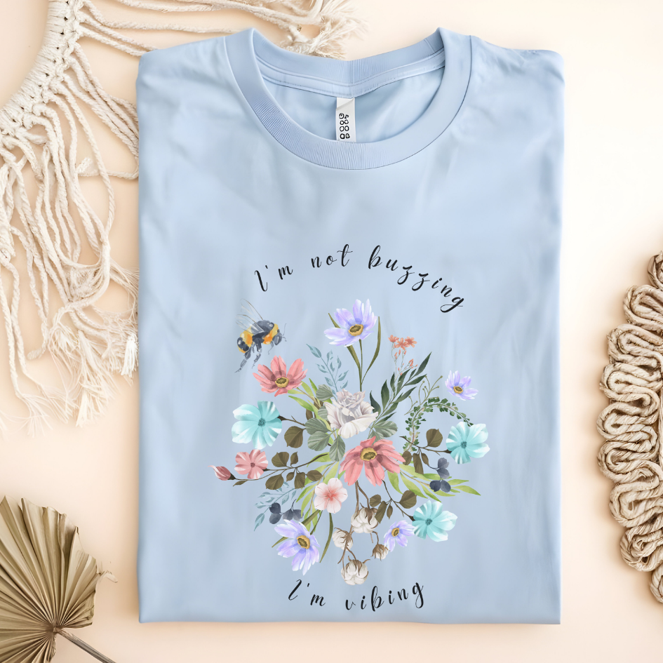 Flat lay of the "Sweet Spring! I'm Not Buzzing" T-Shirt, highlighting the soft, 100% ring-spun cotton fabric, classic crew neck, and the adorable bee and wildflower design. Color Light blue