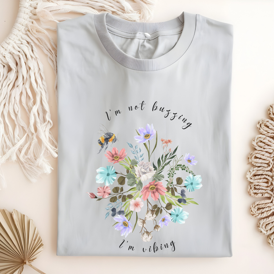 Flat lay of the "Sweet Spring! I'm Not Buzzing" T-Shirt, highlighting the soft, 100% ring-spun cotton fabric, classic crew neck, and the adorable bee and wildflower design. Color Ice grey