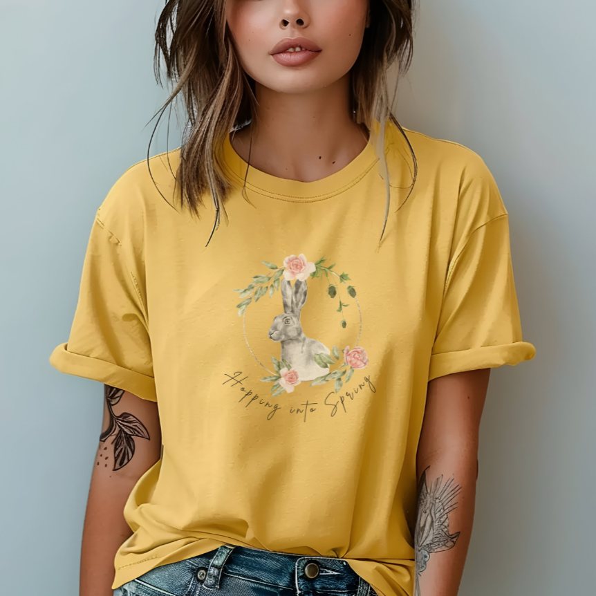 "Hoppin' into Spring" Unisex Garment-Dyed T-Shirt, featuring a delicate watercolor illustration of a grey rabbit framed by a circular wreath of pink roses and lush green foliage, modeled by a Woman wearing size L (oversized). Color Butter