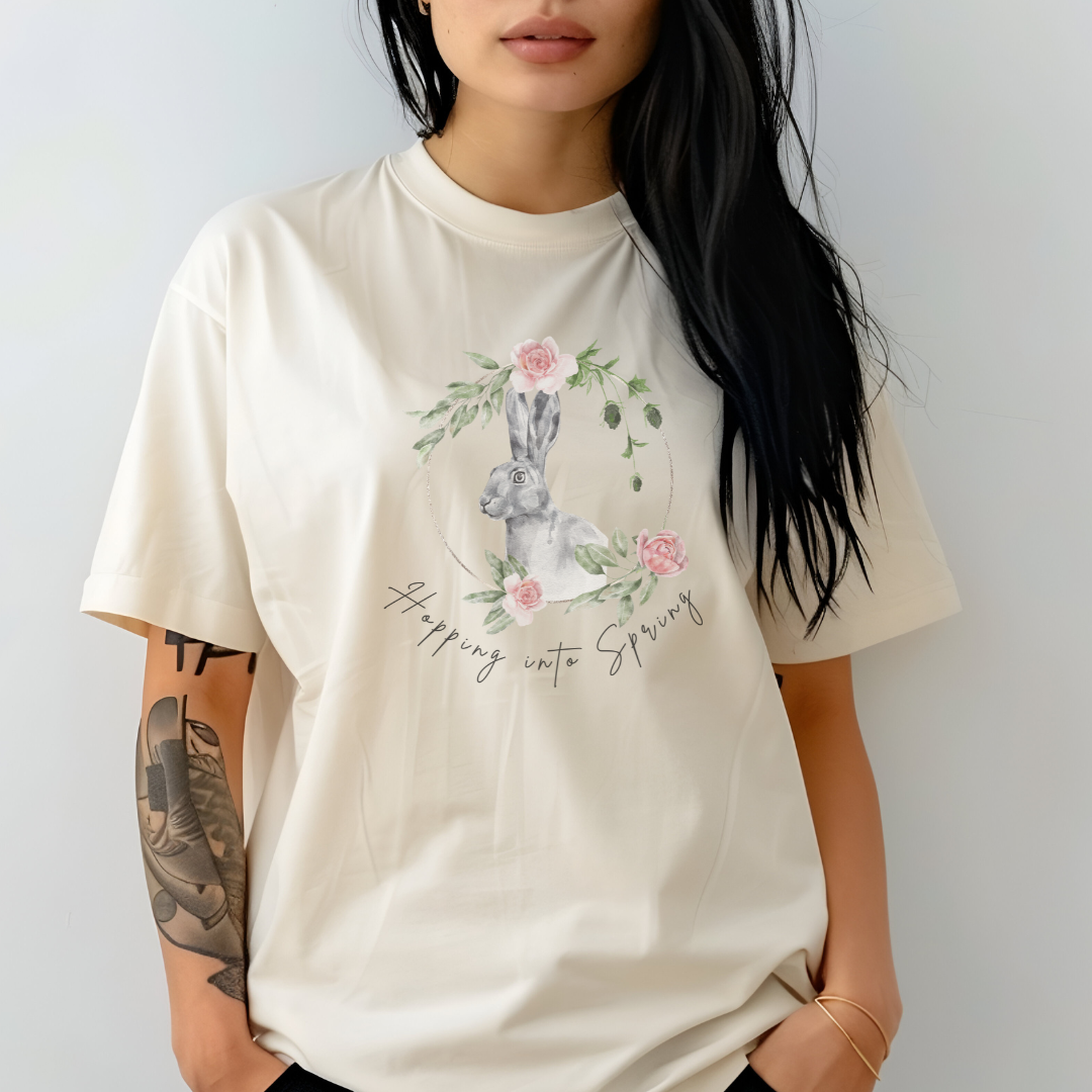 "Hoppin' into Spring" Unisex Garment-Dyed T-Shirt, featuring a delicate watercolor illustration of a grey rabbit framed by a circular wreath of pink roses and lush green foliage, modeled by a Woman wearing size L (oversized). Color Ivory