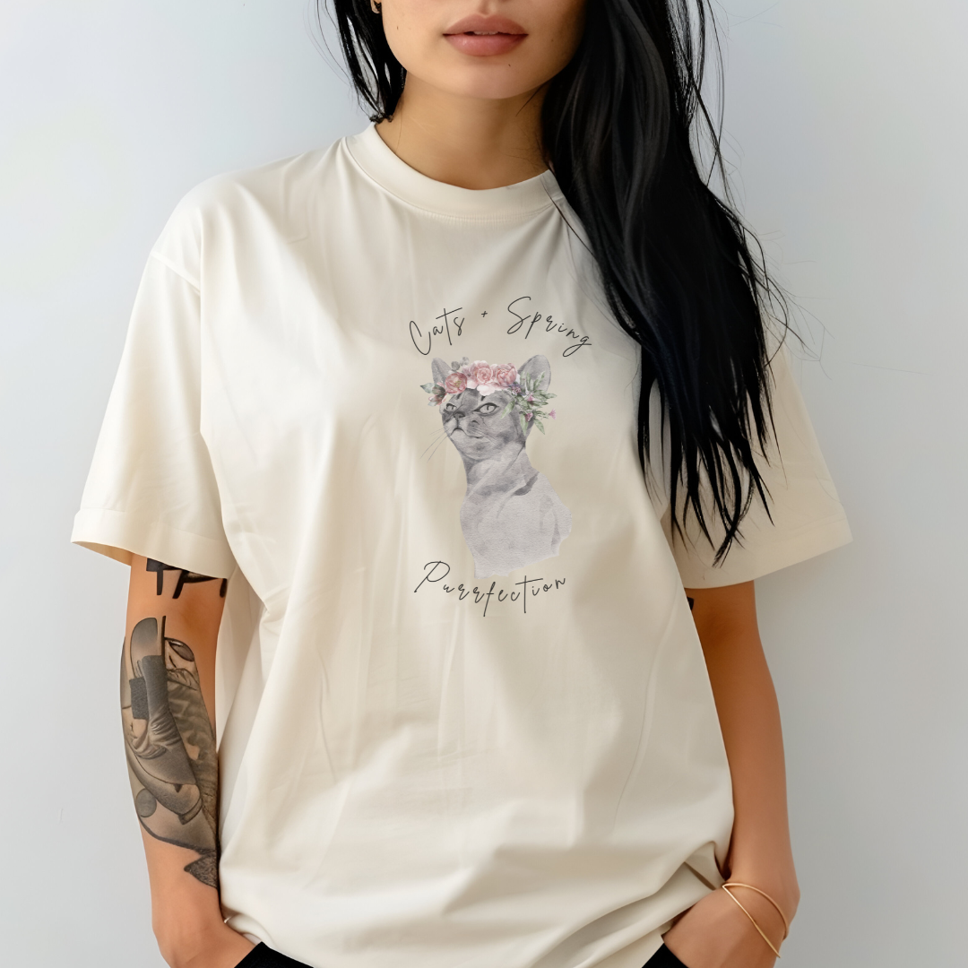 "Purrfect Spring Vibes" Personalized Unisex Garment-Dyed T-Shirt, featuring a charming watercolor illustration of a grey cat wearing a crown of pink roses and green leaves, with the text "Cats + Spring Purrfection" above and below, modeled by a woman wearing size L for an oversized effect. Color Ivory