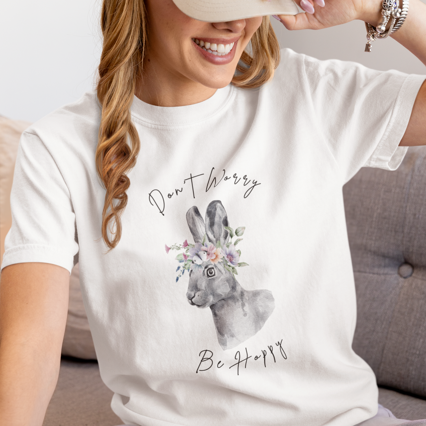 "Don't Worry, Be Hoppy!" Unisex Garment-Dyed T-Shirt, featuring a charming watercolor illustration of a grey rabbit wearing a crown of delicate pink and purple flowers, with the text "Don't Worry Be Hoppy" in a playful script above and below, modeled by a woman wearing size L. Color White