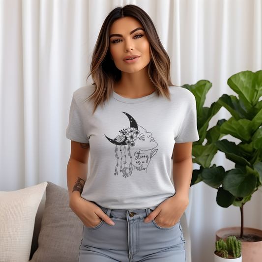 "Strong as a Mother" Unisex Jersey T-Shirt, featuring a powerful graphic of a woman's profile emerging from a crescent moon, adorned with blooming flowers and delicate dangling charms, modeled by a Woman  wearing size M. Color Ash