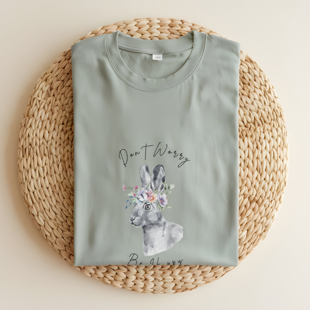 Flat lay of the "Don't Worry, Be Hoppy!" T-Shirt, highlighting the garment-dyed fabric's texture and the durable double-needle stitching. The watercolor bunny and floral crown design are clearly visible. Color White Bay