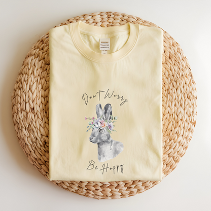 Flat lay of the "Don't Worry, Be Hoppy!" T-Shirt, highlighting the garment-dyed fabric's texture and the durable double-needle stitching. The watercolor bunny and floral crown design are clearly visible. Color Banana