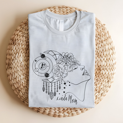 Close-up view of the "Kinda Mom" design on the Mother's Day Tee, showcasing the graphic of a  minimalist line drawing of a woman's profile facing right., her hair is adorned with a crescent moon and dangling charms, giving a mystical or bohemian vibe.  Overlaid on her hair is a cluster of blooming flowers, adding a touch of femininity and nature.  Below the profile, in a casual, handwritten-style font, are the words "Kinda Mom." Color Ash