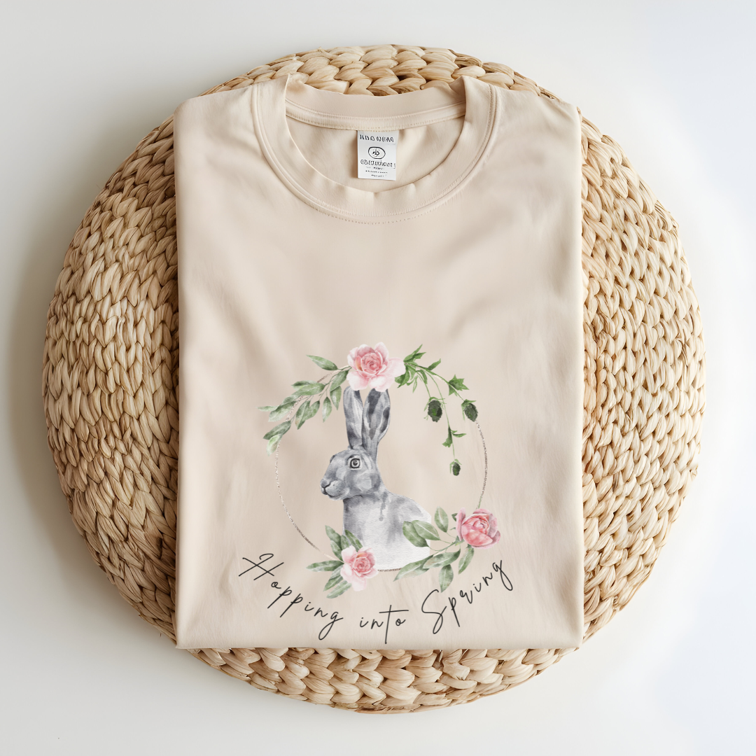 Flat lay of the "Hoppin' into Spring" T-Shirt, highlighting the garment-dyed fabric's texture and the durable double-needle stitching. The watercolor bunny and floral wreath design are clearly visible. Color Ivory