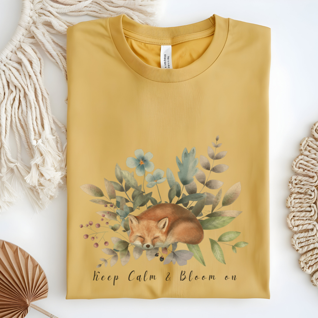 Flat lay of the "Keep Calm & Bloom On" T-Shirt, highlighting the soft, 100% US cotton fabric and classic crew neck. The watercolor fox and floral design are clearly visible. Color Vegas Gold