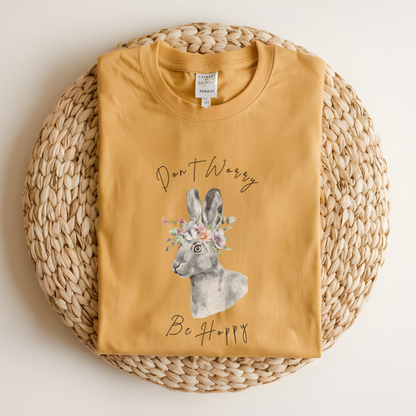 Flat lay of the "Don't Worry, Be Hoppy!" T-Shirt, highlighting the garment-dyed fabric's texture and the durable double-needle stitching. The watercolor bunny and floral crown design are clearly visible. Color Mustard