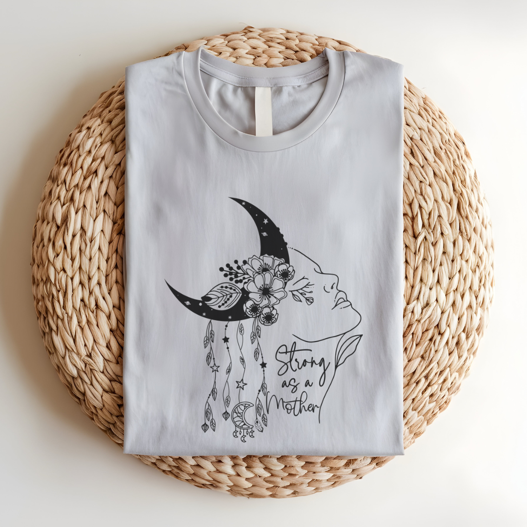Close-up view of the "Strong as a Mother" graphic, showcasing the intricate details of the crescent moon with its stippled texture, the vibrant flowers nestled within it, and the flowing lines of the woman's profile. The dangling charms add a touch of whimsy. The words "Strong as a Mother" are written in a delicate script, font beneath the graphic. Color Heather Clay Grey