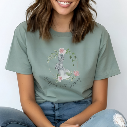 "Hoppin' into Spring" Unisex Garment-Dyed T-Shirt, featuring a delicate watercolor illustration of a grey rabbit framed by a circular wreath of pink roses and lush green foliage, modeled by a Woman wearing size L (oversized). Color Bay